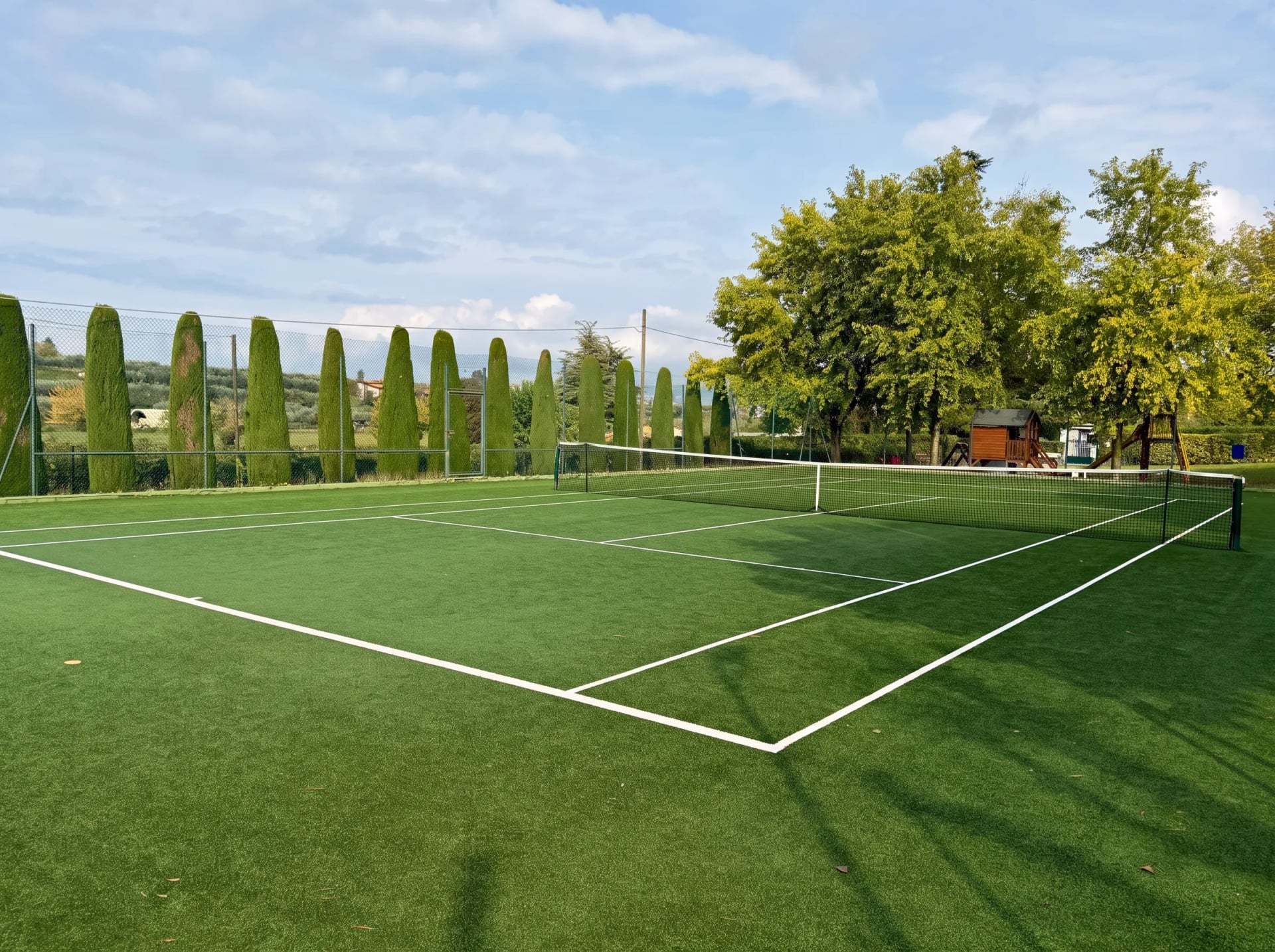 Tennisplatz mit grünem Kunstrasen, dahinter eine Reihe Zypressen und Bäume